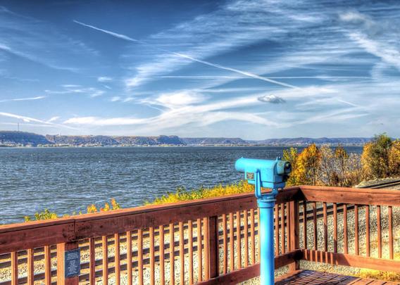 A scenic landscape features a telescopic viewer on a wooden deck overlooking the Mississippi River and distant hills under a sky filled with wispy clouds.