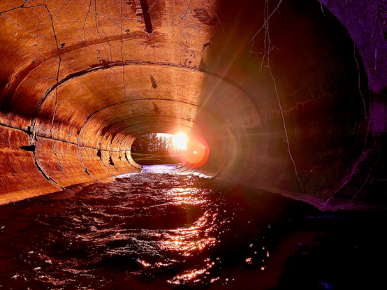 Color image of the moment the setting sun aligns with the culvert/sewer pipe conducting Yellow Creek under a road in a suburban neighborhood, Bath Township, Summit County, Ohio, USA. Image shows the sun directing its beams through the culvert from end to end, lighting up the creek surface and setting the walls of the culvert aglow in shades of orange, yellow, and red.