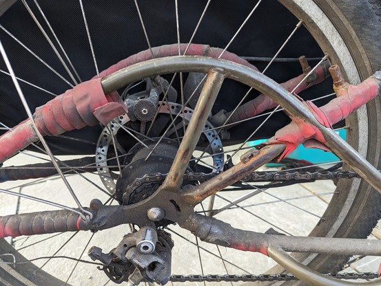 The rear frame and wheel of a venerable Big Dummy with the cargo carrying sides removed.  The frame was previously wrapped with squishy red handlebar tape to protect it from cargo/bag abrasion, but some of that has worn away or come loose, exposing tubes, some with the paint worn away to bare metal, and much rust around the exposure.  The hub (large, block, Rohloff 14-speed) is filthy with lubricant and road dust residue, the frame near the hub is similarly vile, the derailer-repurposed-as-chai…