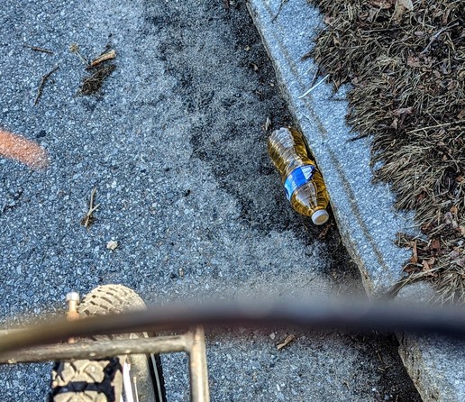 A water bottle in the street next to a curb.  The water is full of yellow liquid.  Just because The phone was on my bike, there's brake cables, front rack, and bike tire on the bottom edge of the photo and in the blurred foreground.