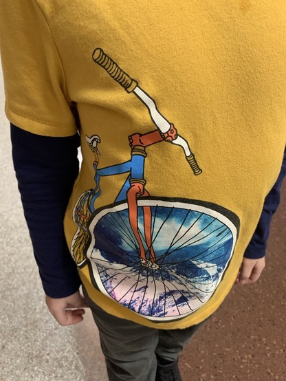 A blue and orange bicycle graphic on a child’s goldenrod shirt. A scene through the front spokes looks like a snow capped mountain top with a blue sky and white clouds above. 