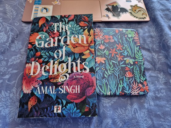 A book - The Garden of Delights by Amal Singh - and a covered Kindle side-by-side in front of a laptop on a bed. Both have dark floral designs.