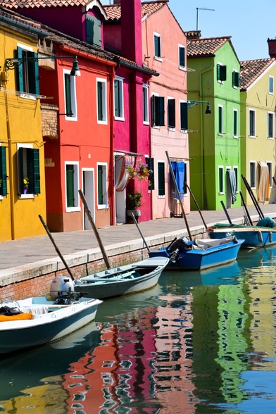 A row of houses along a canal in Italy. Their bright colors of mustard , orange, pink green and light yellow reflect in the rippled water of the canal. Four small open boats with outboard motors are moored along the canal’s edge.