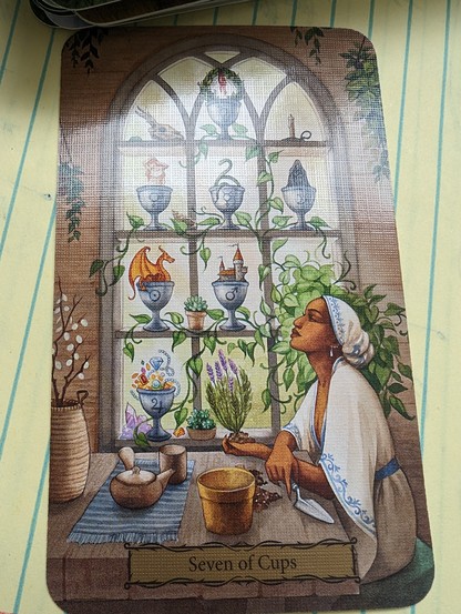 Seven of Cups. A black woman with a white head wrap and white dress is seated at a wooden table in front of a window shelf that holds 7 cups of possibility.  She has a trowel in her right hand and a purple flower (lavender?) ready to plant in her left.