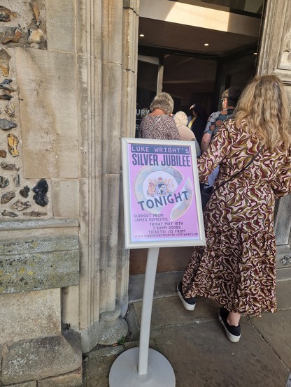 A queue of people entering a demonstrated church with a poster for Luke Wright's Silver Jubilee outside