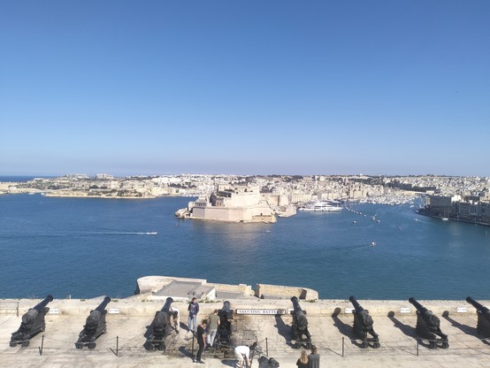 View out across Valletta harbour from the salting battery to the three  fortified cities, of Vittoriosa, Senglea and Cospicua in the distance. Sky is blue and clear, sea is blue and calm.