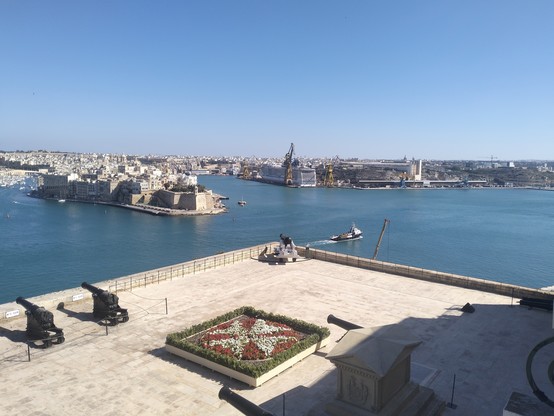 View out across the harbour, a flower bed planted up with white flowers in a Maltese cross, with red flowers around the edges, is in the foreground. Cannons face out to the harbour, the 3 cities of Vittoriosa, Senglea and Cospicua are in the distance, and on the left the cruise liner dry dock where a behemoth of a vessel undergoes a service.