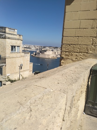 View over the Valletta fortifications into the harbour, a boat motors from left to right through the deep blue water. The strong limestone tones contrast to the bright blue sky and the deep blue of the Mediterranean sea.