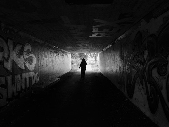 Black and white photograph of a figure walking towards the end of a tunnel. The photo is taken from the inside of the tunnel, with the walls being covered in graffiti and only being illuminated by the sunlight coming from outside the tunnel.