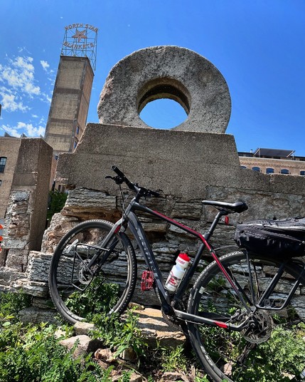 A black bicycle leans against a cement wall with a grind stone on top of it. Beyond is a tall tower structure with an old sign that says ‘North Star Blankets’ There is a blue sky above with a few wispy clouds next to the tower.