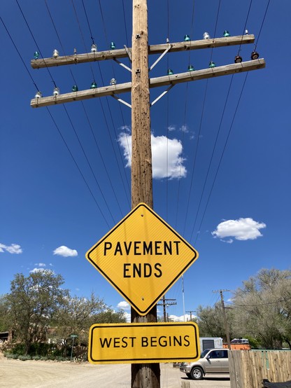 Photo of a road sign saying “Pavement Ends” with a sign below saying West Begins” on an electric pole with some beautiful glass insulators.
