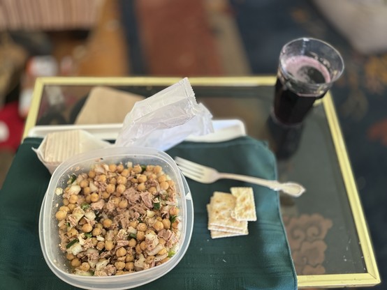 A container with chickpea and tuna meat salad, a stack of crackers, a plastic-wrapped utensil, a folded napkin, and a glass of grape juice on a table.