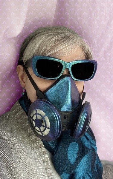 Person with gray hair wearing teal iridescent sunglasses and a matching respirator mask with a silver cardigan