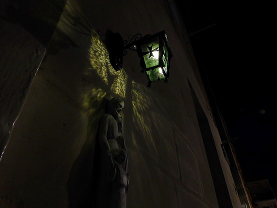 Night photograph of a lantern shedding a soft greenish light on the limestone wall that it is attached to. Under the lantern is a small stone statue of a man, a woman and a child. The lantern is decorated with the Maltese cross, and the shadow from the cross can be seen cast on the wall, above the heads of the statue.
