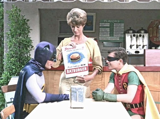 Batman and Robin sit at a table while a waitress shows them a menu advertising a 