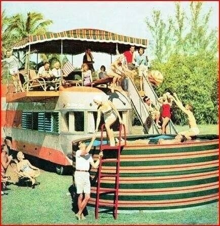 A group of people enjoying a sunny day around a retro bus conversion with a slide leading into a large, colorful above-ground pool.
