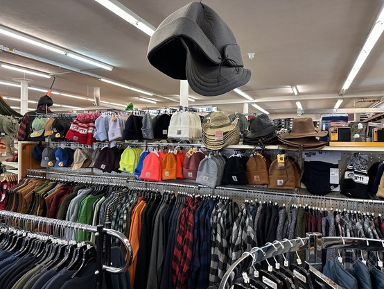 Under thin strips of fluorescent lights on the ceiling are racks of flannels and wool shirts. Above one line tie of clothes are two rows of hanging hats. Above this is a giant Mackinaw Wool cap. 