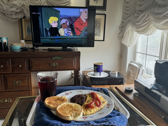 Plate of food with English muffins, an omelet, and ketchup in the foreground, a glass of red drink, a TV showing an animated cartoon in the background, and a cluttered room with dresser and electronic equipment.