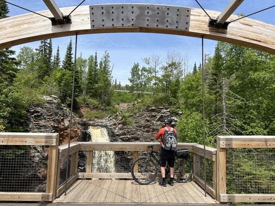 A person in an orange jacket and black bicycle stands next to a black bicycle. He is on a wooden bridge looking at a waterfall and taking a photo of it. There is blue sky above and the bridge arched above his head.