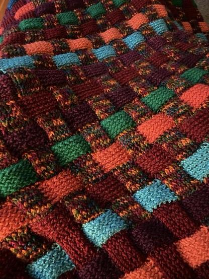 A colorful knit blanket featuring an interlocking pattern with various shades of green, orange, red, blue, and multicolor yarn.