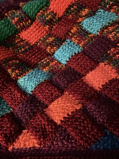 A colorful knitted pattern featuring interwoven squares in shades of red, orange, blue, green, and multicolor threads.
