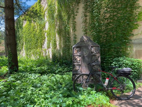 In a lush, green scene a black bicycle leans on a stone monument to a natural history museum.