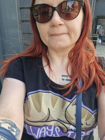 Selfie of a woman with red hair, wearing sunglasses and a top that says 'always tired' with a picture of a cat.