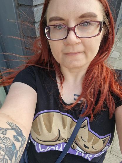Selfie of a woman with red hair, wearing regular glasses and a top that says 'always tired' with a picture of a cat.