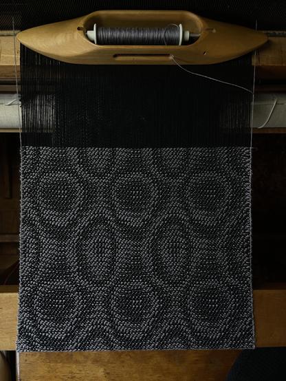 Photo of weaving in progress on a loom taken from directly above. The pattern is irregular circles and ovals in black and grey. 