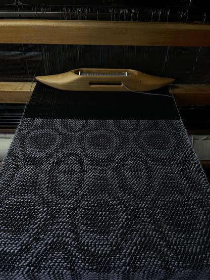 Photo of weaving on the loom. The pattern is irregular circles and ovals in black and white. The photo is taken from a forward angle and accentuates the length of the shapes. 