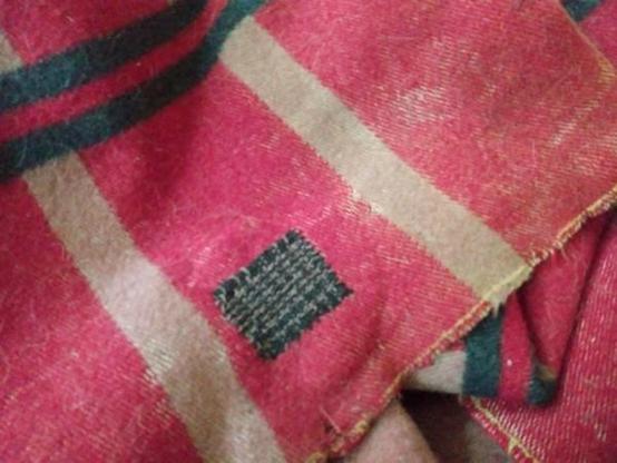A threadbare red blanket with beige stripes with a square darning patch of black and beige woven threads. 