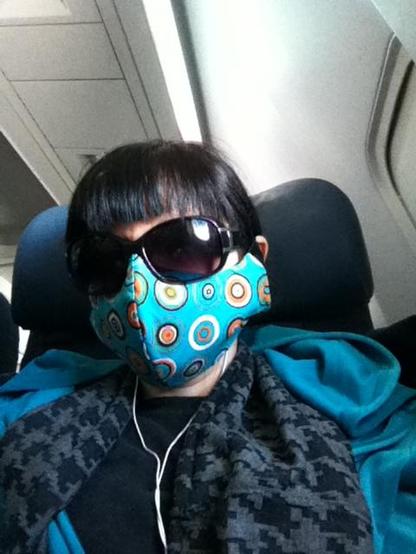 Person with dark hair in airplane seat wearing oversized sunglasses, earbuds, a scarf, teal hoodie and a fabric, teal patterned mask.