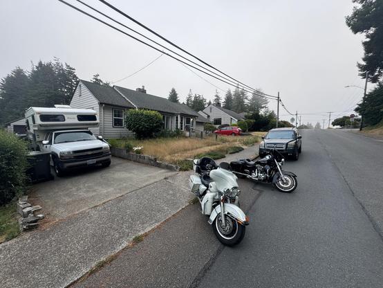 A residential street with houses, a white truck with a camper in the driveway, a red car, a black SUV, and two motorcycles parked on the road. Overcast sky and trees in the background.
