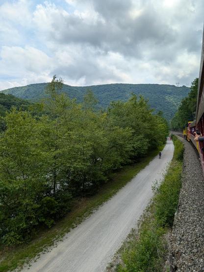 A train travels through a mountain pass on a curve which is going to the left. Left there is a large row of trees on the left and between the tracks and the trees is a bike path with several cyclists on it. In the background is tree covered hill and the sky is dark and stormy