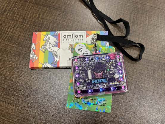 A HOPE attendee badge with flashing blue and purple LEDs, he/him and they/them pronoun stickers and an omnom-branded, rainbow-striped chocolate bar with abstract white unicorns printed on it lie on a dark brown tabletop. 