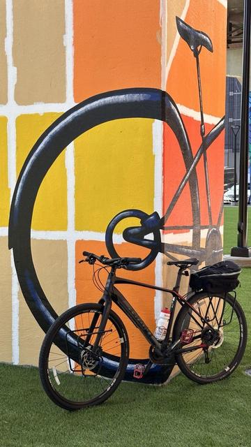 A black bicycle next to a large painting of a black bicycle on a bridge support pillar. The background of the painting has large yellow, tan, and orange squares.