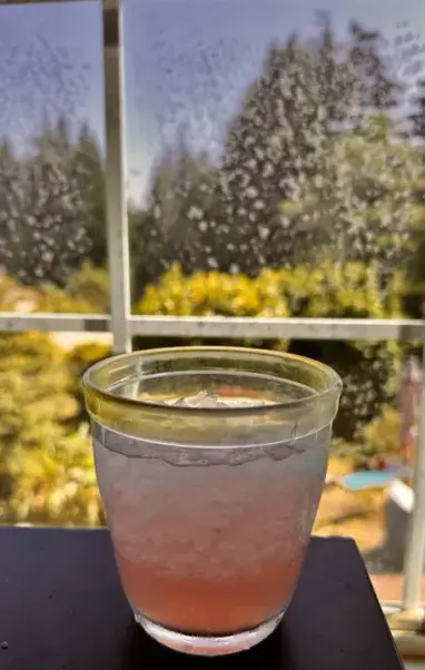 A pink cocktail with lots of ice with a view out a window showing greenery
