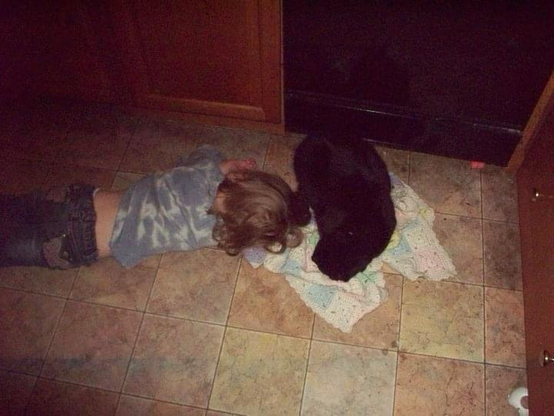 Toddler sleeping on the floor with her head on a crocheted blanket that has a black cat laying on it. They are laying in front of a dishwasher.