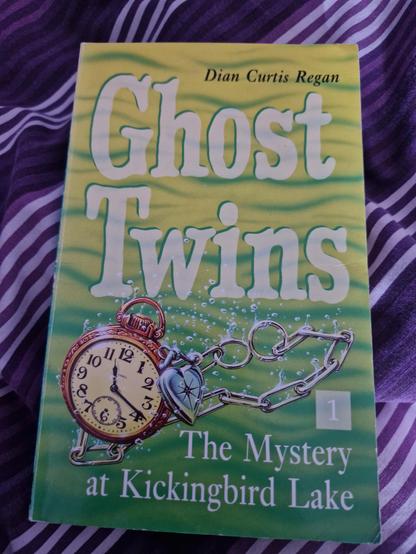 Paperback of Ghost Twins 1: The Mystery at Kickingbird Lake by Dyan Curtis Regan, on a purple and white striped duvet cover. The book is coloured in green and yellow waves and features a pocketwatch and a heart-shaped locket on a chain