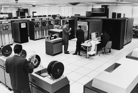 A room full of racks of equipment including large tape machines. Several people can be seen working on the equipment