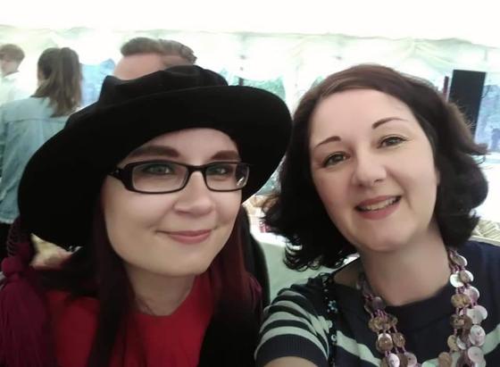 Two women posing for a selfie in a marquee. The one on the left has red hair and glasses, and is dressed in a PhD gown and hat. The one on the right has dark hair and is wearing a stripy top and necklace made of pink shells.