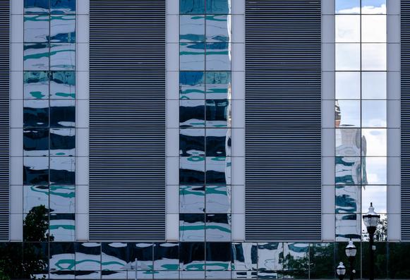 the side of a glass curtain building, featuring repeated squares of window panels (each reflecting aspects of the surroundings) and some large louvered areas seen as horizontal stripes; there are also three street lamp tops in a repeating series in a sequence of ascending/descending apparent sizes