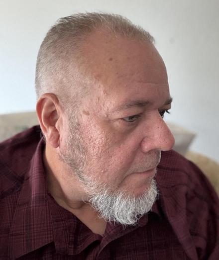 A close-up side profile of an older man with short hair, a trimmed white beard, and wearing a maroon checkered shirt.
