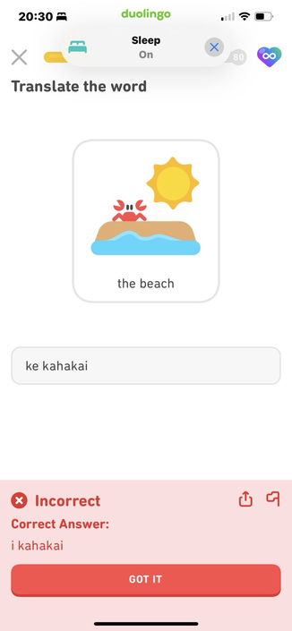 Screenshot from Duolingo. The lesson prompt is a cartoon image of a tan horizontal area representing sand with a wavy blue area beneath representing waves; a red cartoon crab stands atop the sand, and a yellow cartoon sun hangs above. Lesson prompt text: “the beach.” I have answered “ke kahakai” but at the bottom is the judgement “Incorrect! Correct answer: i kahakai” in a bright red box!