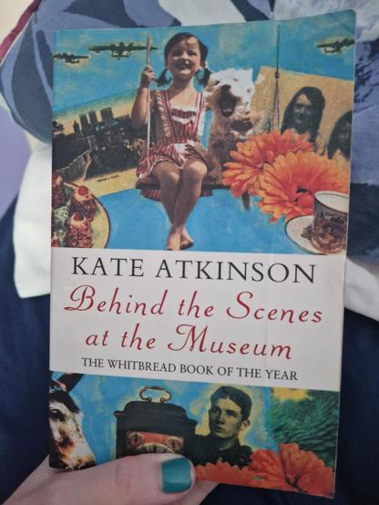 Hand holding up a paperback of Behind the Scenes at the Museum by Kate Atkonson, which features a collage of photos including a girl on a swing, a horse, mid-20th century buffet food, WWII planes and soldier, and orange zinnias