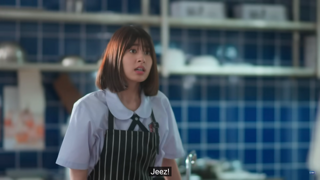 Mudmee stands in a blue-tiled public kitchen for the school. She wears a light blue button down shirt with a wide, rounded collar, and an apron with pinstripes. She looks panicked and annoyed in equal measure.

The captions says: 