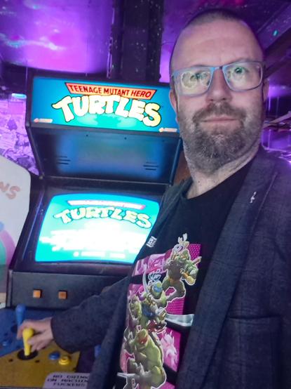 Game developer Steven Goodwin wearing a shirt for the new TMNT game, standing in front of an arcade cabinet, or the old TMNT game