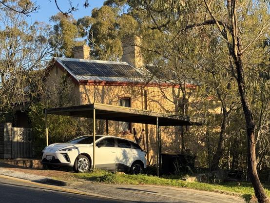 Old stone cottage with solar panels and electric car parked out front, with trees, mostly eucalypts