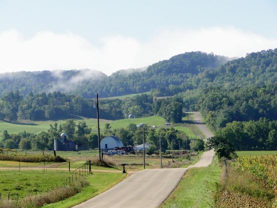 Foto mit maximaler Zoomeinstellung des Teleobjektivs. Blick nach Süden, ein heißer Sommertag, Wolken steigen vom Bergkamm auf. Links eine Milchfarm, eine kleine Straße steigt und fällt über das Land, das in die Hügel aufsteigt. Rechts ein bisschen Mais, Wiesen, wo es eben genug ist.