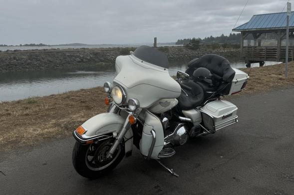 A white motorcycle parked by a waterfront on a cloudy day. A picnic shelter with a blue roof is in the background.